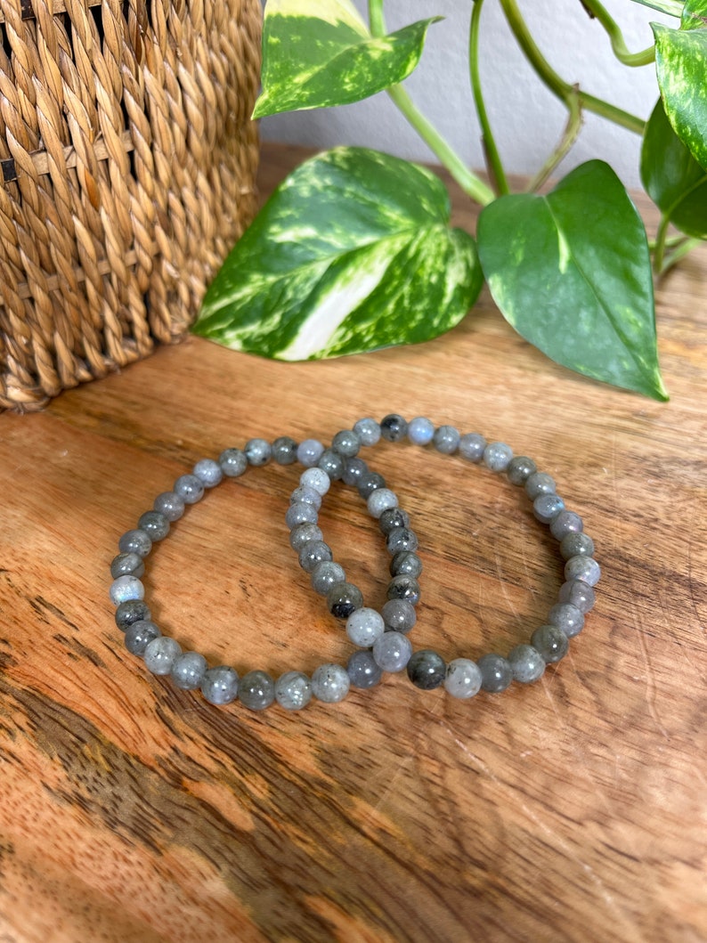 May include: Two grey beaded bracelets made of labradorite stones. The bracelets are stacked on top of each other on a wooden surface.