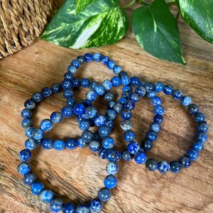 May include: Four blue lapis lazuli beaded bracelets stacked on a wooden surface. The beads are round and have a speckled pattern.