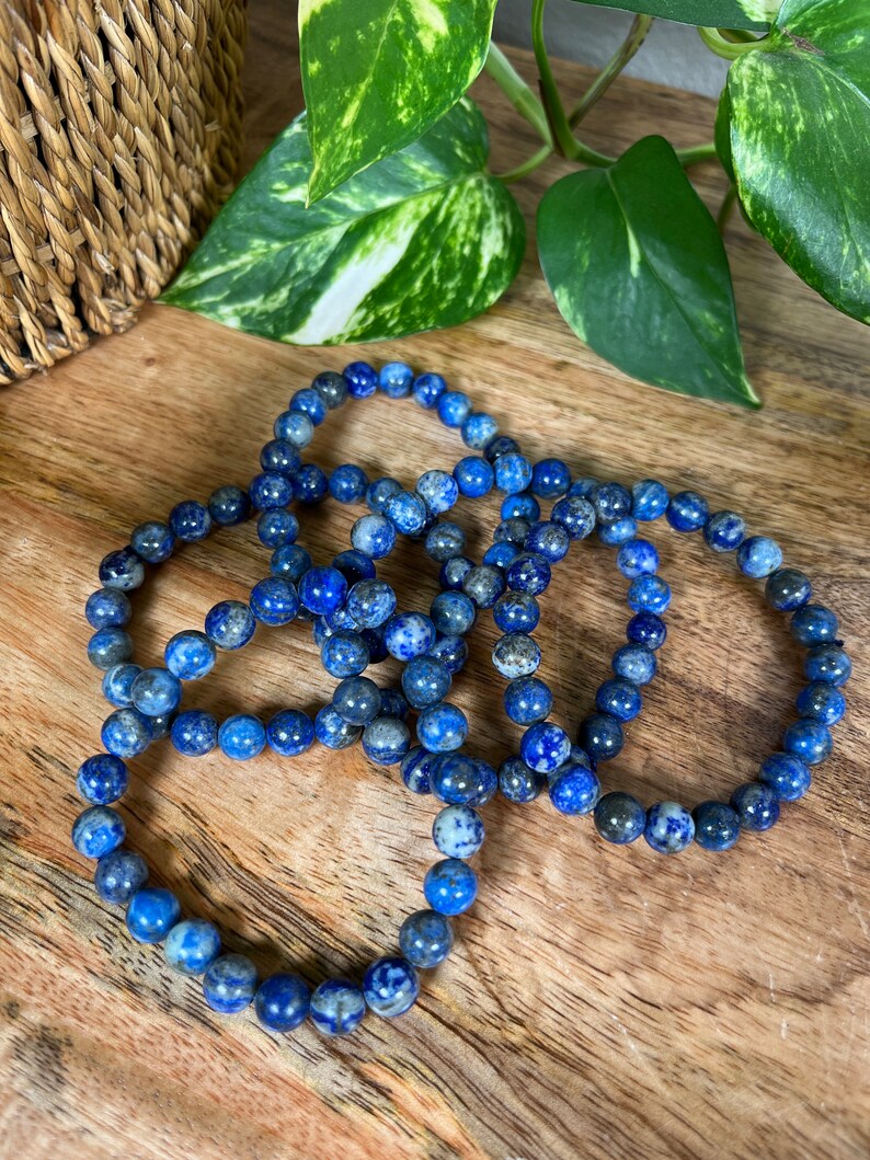 May include: Four blue lapis lazuli beaded bracelets stacked on a wooden surface. The beads are round and have a speckled pattern.