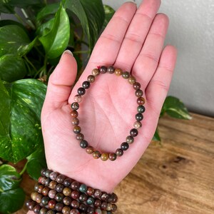 May include: A brown, green, and red gemstone bracelet with small round beads. The bracelet is on a person's hand.