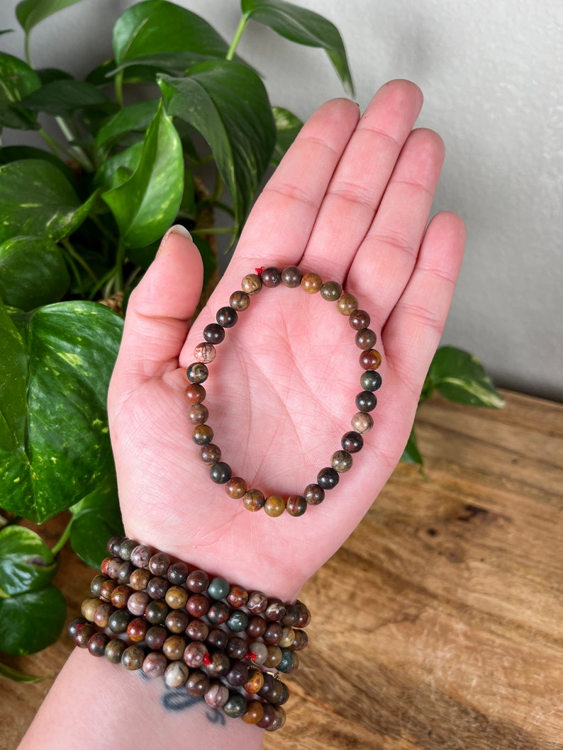 May include: A brown, green, and red gemstone bracelet with small round beads. The bracelet is on a person's hand.
