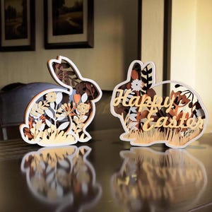 Puede incluir: Dos decoraciones de conejito de Pascua de madera con intrincados diseños florales. El conejito más grande tiene las palabras "Happy Easter" cortadas en su cuerpo.