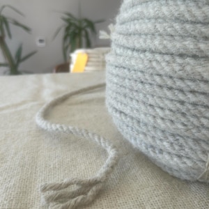 May include: A large spool of light grey rope with a twisted design. A length of the rope extends from the spool, with the end frayed. The rope is on a light-coloured, textured surface. Plants are visible in the background.