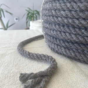 May include: A grey rope spool on a white surface. The rope is tightly wound, with a length extending from the spool. The rope has a twisted, braided texture. The background is blurred, with hints of plants.