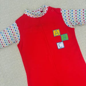 Puede incluir: Mono de beb&eacute; rojo con mangas largas y un patr&oacute;n de lunares blancos y coloridos en las mangas. El mono tiene un cuello alto y una cremallera. La parte delantera del mono tiene un dise&ntilde;o bordado de colores de las letras A, B y C en bloques.