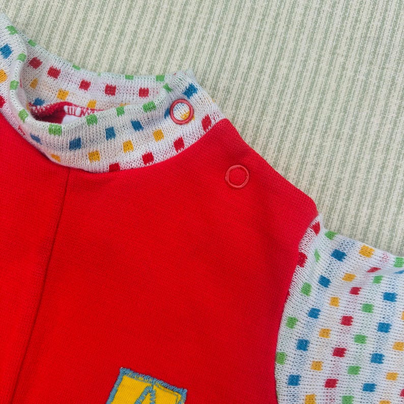 Puede incluir: Mono rojo de manga larga con un estampado de cuadros coloridos alrededor del cuello y las mangas. El mono tiene un peque&ntilde;o parche amarillo con una letra A en la parte delantera.