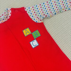 Puede incluir: Un pelele rojo para beb&eacute; con mangas largas y un estampado de lunares de colores en las mangas. El pelele tiene una cremallera y un parche con las letras A, B y C en bloques de colores.