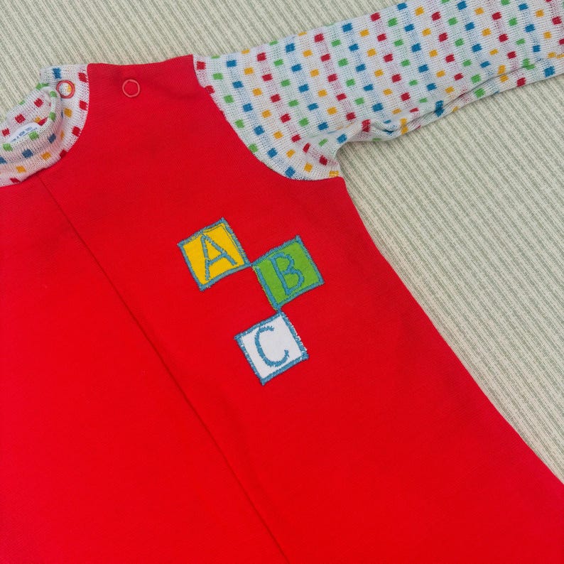 Puede incluir: Un pelele rojo para beb&eacute; con mangas largas y un estampado de lunares de colores en las mangas. El pelele tiene una cremallera y un parche con las letras A, B y C en bloques de colores.