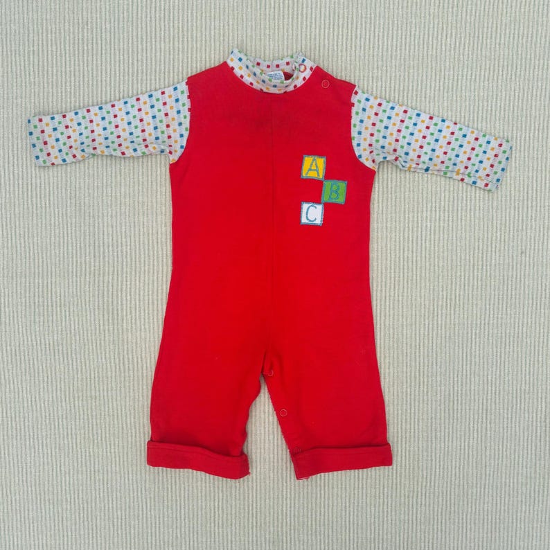 Puede incluir: Mono de beb&eacute; rojo con mangas largas y un patr&oacute;n de lunares blancos y coloridos en las mangas. El mono tiene un cierre de presi&oacute;n en la entrepierna y un parche con las letras A, B y C en la parte delantera.
