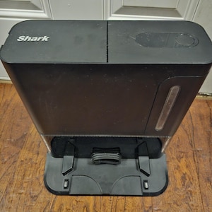May include: A black Shark robot vacuum charging station. The unit has a rectangular shape with a divided top and a base with charging contacts. The Shark logo is visible on the top.