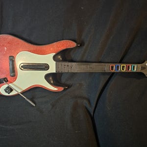 May include: A red and white plastic guitar controller with a black neck and colorful fret buttons. The guitar has a whammy bar and a white pickguard. The body of the guitar is red with a worn appearance.