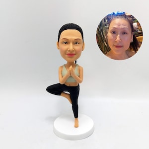 May include: A custom figurine of a person in a yoga pose. The figurine is made of painted ceramic and has a realistic appearance. The person is wearing a light green tank top and black leggings. The figurine is standing on a white base.