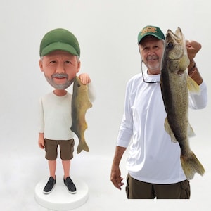 May include: A custom bobblehead figurine of a person wearing a green baseball cap, white shirt, brown shorts, and black shoes. The person is holding a fish in their right hand. The bobblehead is standing on a white base.