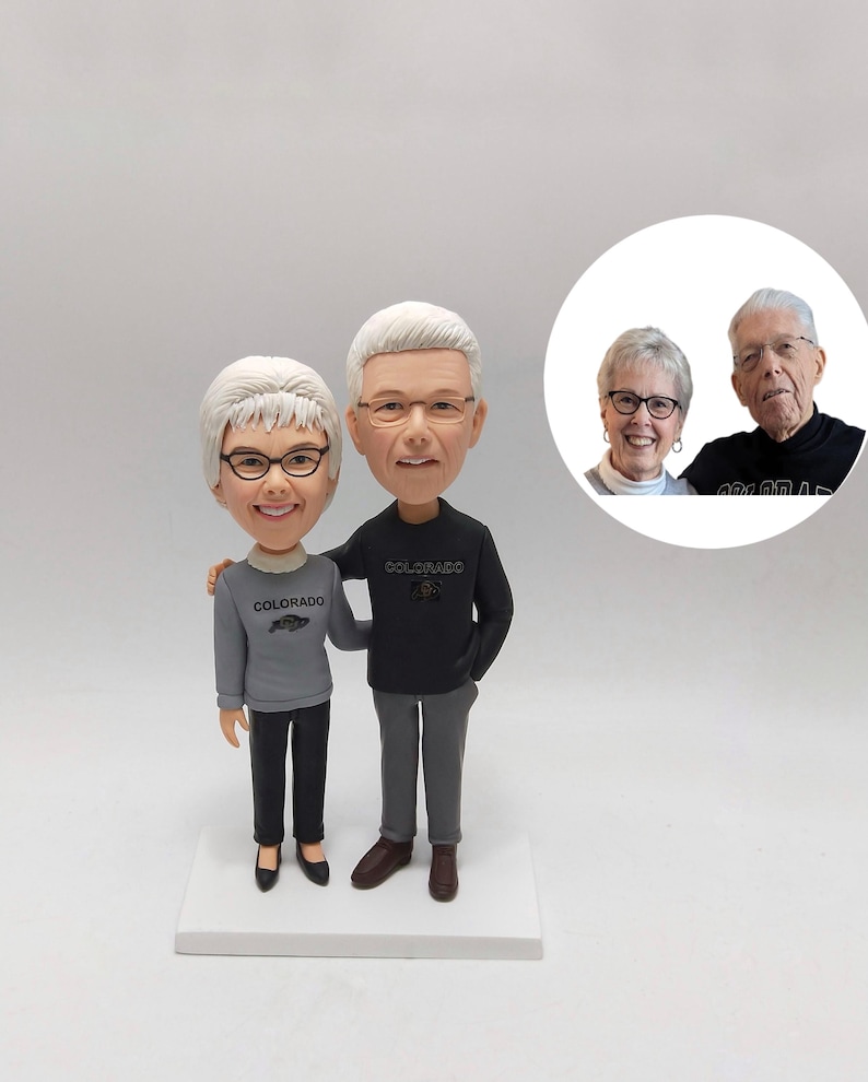 May include: A personalised bobblehead figurine of a couple, the woman in a grey sweatshirt with "Colorado" printed on it, and the man in a black sweatshirt with "Colorado" printed on it. The figures stand on a white base.
