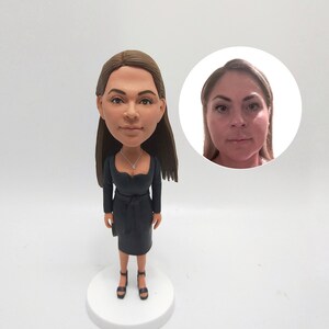 May include: A custom-made figurine of a person wearing a black dress and black shoes. The figurine is standing on a white base. A close-up photo of the person is shown in a circle behind the figurine.