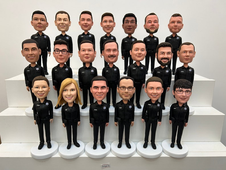 May include: A group of bobblehead figurines depicting people in dark uniforms, standing on white circular bases. The figurines have detailed faces and are arranged on a tiered white display. The uniforms have a badge on the chest.