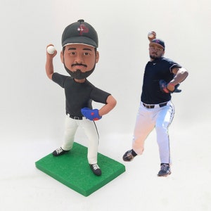 Könnte beinhalten: Eine personalisierte Baseball-Figur eines Werfers in einem schwarzen Trikot, einer weißen Hose und einer schwarzen Kappe mit einem roten Logo. Die Figur hält einen weißen Baseball und steht auf einem grünen Sockel. Die Figur ist in einer Wurfposition dargestellt.