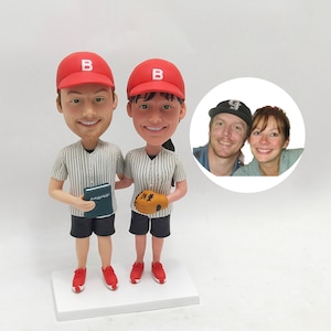 May include: A custom-made figurine set of a couple dressed in baseball uniforms. The figurines are wearing red baseball caps with a white "B" on the front. The man is holding a book and the woman is holding a baseball glove. A photo of the couple is included in the background.