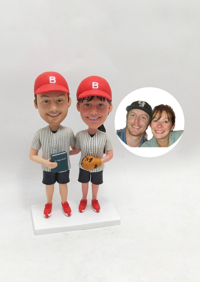 May include: A custom-made figurine set of a couple dressed in baseball uniforms. The figurines are wearing red baseball caps with a white "B" on the front. The man is holding a book and the woman is holding a baseball glove. A photo of the couple is included in the background.