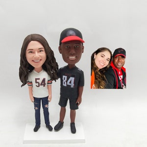 May include: A custom-made figurine set of a couple wearing sports jerseys. The woman is wearing a white jersey with the number 54 and blue jeans. The man is wearing a black jersey with the number 84 and black shorts. The figurines are standing on a white base.