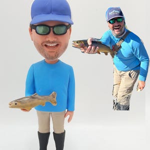 May include: A blue and white custom bobblehead figurine of a person wearing a blue hat, sunglasses, and a blue shirt, holding a fish. The figurine is standing on a white base.