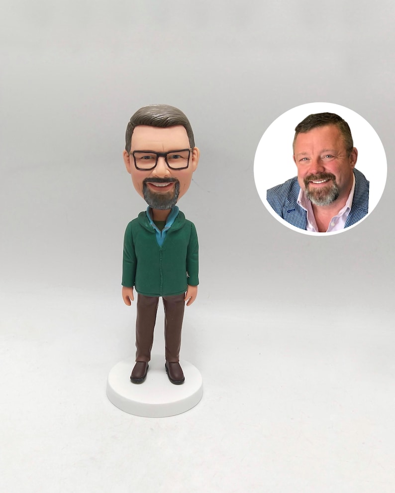 May include: A custom bobblehead figure of a man wearing glasses, a green hoodie, and brown trousers. The figurine stands on a white base. A circular image of the man is next to the figure.