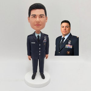 May include: A custom bobblehead figurine of a person wearing a dark blue military uniform with a light blue shirt and a dark tie. The figurine is standing on a white base. The person's face is depicted in a cartoon style.