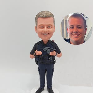 May include: A custom-made figurine of a police officer wearing a blue uniform with a black belt and a black vest. The figurine is standing on a white base. The officer is holding a radio and a flashlight. The figurine is a realistic depiction of a police officer.