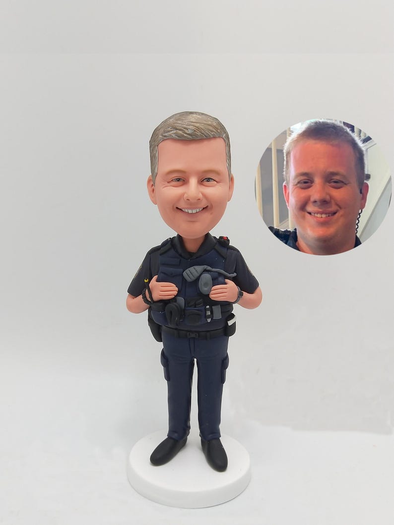 May include: A custom-made figurine of a police officer wearing a blue uniform with a black belt and a black vest. The figurine is standing on a white base. The officer is holding a radio and a flashlight. The figurine is a realistic depiction of a police officer.