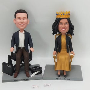 May include: Two custom bobbleheads, one of a person in a black suit sitting on a director's chair and the other of a person wearing a gold dress and a crown sitting on a throne. The bobbleheads are on a gray base with the text "Director of the West" and "Queen of the East" printed on them.