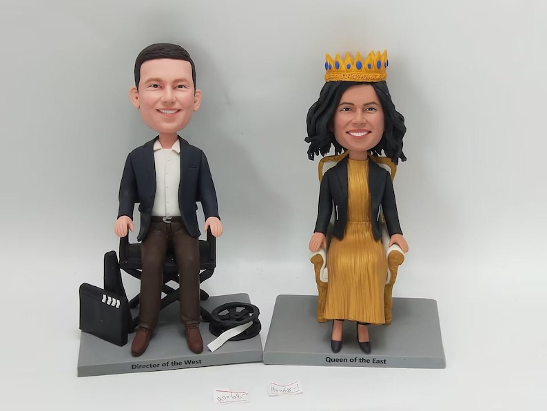 May include: Two custom bobbleheads, one of a person in a black suit sitting on a director's chair and the other of a person wearing a gold dress and a crown sitting on a throne. The bobbleheads are on a gray base with the text "Director of the West" and "Queen of the East" printed on them.