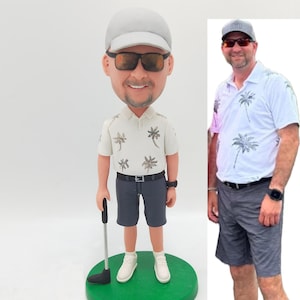 May include: A custom bobblehead figurine of a person wearing a white hat, sunglasses, a white shirt with palm trees, gray shorts, and white sneakers. The figurine is holding a golf club and is standing on a green base. The person is also pictured in a separate image wearing a similar outfit.
