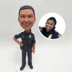 May include: A bobblehead figurine of a smiling police officer in a dark blue uniform, standing on a white base. A smaller image of a smiling woman in a police uniform is in the background.