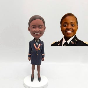 May include: A custom bobblehead figurine of a person wearing a dark blue uniform with a red, white, and blue ribbon and gold buttons. The figurine is standing on a white base.