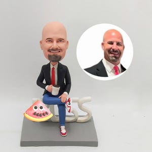 May include: A figurine of a bald man in a black suit and red tie, seated with a coffee cup. He's wearing blue jeans and red sneakers. A slice of watermelon and the letters "SBS" are also present. A smaller image of the man is in the background.