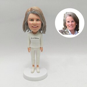 May include: A bobblehead figurine of a woman in a white outfit and trainers. The figurine has a smiling face and grey hair. The text "60 and fabulous" is printed on the shirt. A smaller image of the woman is in the background.