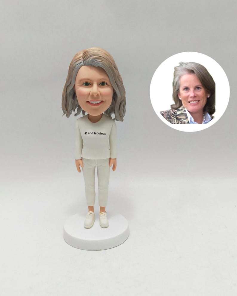 May include: A bobblehead figurine of a woman in a white outfit and trainers. The figurine has a smiling face and grey hair. The text "60 and fabulous" is printed on the shirt. A smaller image of the woman is in the background.