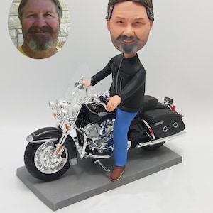 Può includere: Una statuetta personalizzata a testa oscillante di un uomo che guida una motocicletta nera. L'uomo indossa una giacca di pelle nera e jeans blu. La motocicletta ha un parabrezza cromato e dettagli cromati.