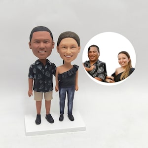 May include: A custom-made figurine set of a couple, with the man wearing a black and white patterned shirt and khaki shorts, and the woman wearing a black top and blue jeans. The figurines are standing on a white base.