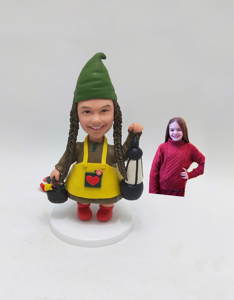 May include: A custom-made gnome figurine with a green hat, brown hair, and a yellow apron with a red heart. The gnome is holding a lantern and a basket of flowers.