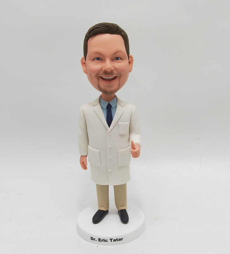 May include: A white doctor's coat with a blue tie and a thumbs up. The figurine is standing on a white base with the text "Dr. Eric Tatar" printed on it.