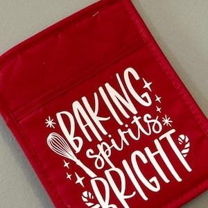 May include: A red oven mitt with the words "BAKING spirits BRIGHT" in white script. The design includes a whisk, stars, and candy cane accents. The mitt has a pocket and a loop for hanging.