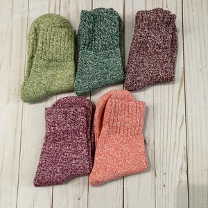May include: Five pairs of cozy socks in various colors are arranged on a light wood surface. The socks include shades of green, burgundy, and pink. The socks have a ribbed cuff and a textured knit design, perfect for cold weather.