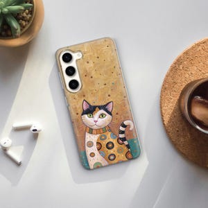Może przedstawiać: Przezroczyste etui na telefon z kolorową ilustracją kota w czarno-białe paski. Kot nosi wzorzysty kołnierzyk i ma złote tło z małymi brązowymi kropkami. Etui ma wycięcie na aparat z trzema obiektywami.