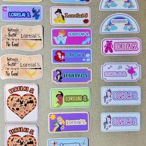 May include: A collection of colorful name tag stickers featuring various designs and the name "Lorelai S." Designs include cartoon characters, rainbows, unicorns, and themed elements from popular stories. The stickers are rectangular and arched.