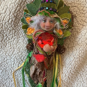 May include: A handcrafted fairy figurine featuring a pointed green hat with colorful sequins, white hair, and a large faceted red gem held in its hands. The figure is adorned with green fabric leaves, small pinecones, and long yellow and green ribbons.