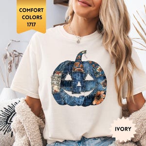 May include: Ivory-colored t-shirt featuring a denim patchwork pumpkin design with a smiling face and floral accents. The shirt has the text "COMFORT COLORS 1717" and "IVORY" on it.