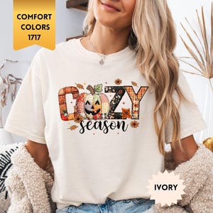 Op de afbeelding: Ivoor kleurig T-shirt met een "Cozy Season" afbeelding met een pompoen en herfstbladeren. Het shirt is een Comfort Colors 1717 stijl. Het model draagt een spijkerbroek short en een crème kleurig vest.
