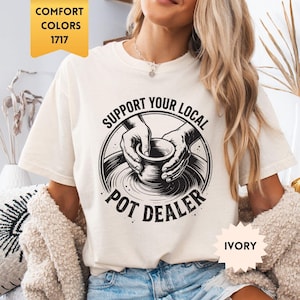 May include: Ivory-colored t-shirt with the text "SUPPORT YOUR LOCAL POT DEALER" in a black circular design. The shirt is a Comfort Colors 1717 style. The model is wearing blue denim shorts and a cream-colored cardigan.