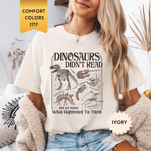 May include: Ivory-colored t-shirt with the text "DINOSAURS DIDN'T READ" and dinosaur illustrations. The shirt also includes the phrase "and we know What Happened To Them". The shirt is a comfort color, size 1717.